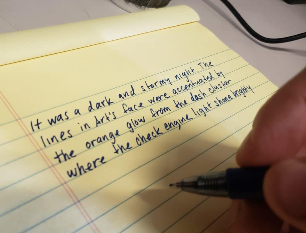 Picture of hand, pen, and paper with writing: "It was a dark and stormy night. The lines in Art's face were accentuated by the orange glow from the dash cluster where the check engine light shone brightly."