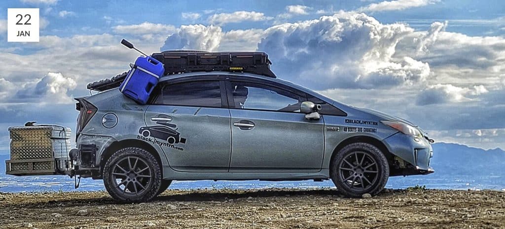 A Prius with a lift kit, roof rack, hitch mounted box, solar panels, and oversized tires on a hill with beautiful sky as a backdrop.