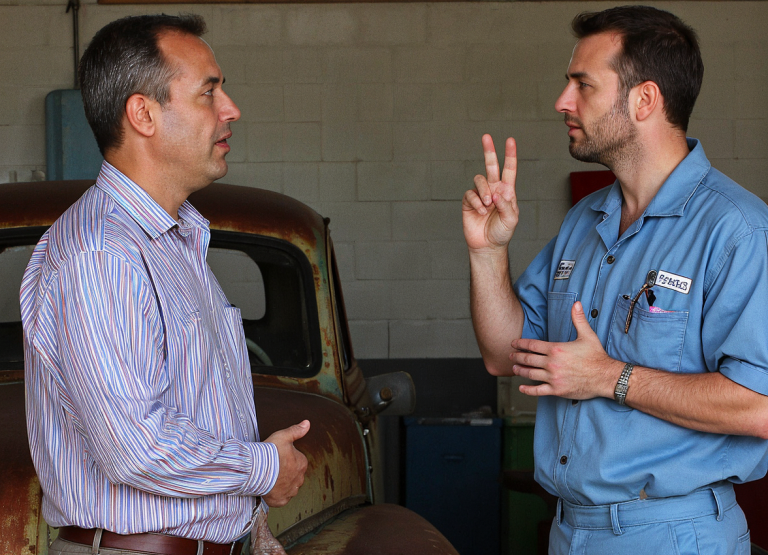 A mechanic talking to a customer in front of a rusty car.